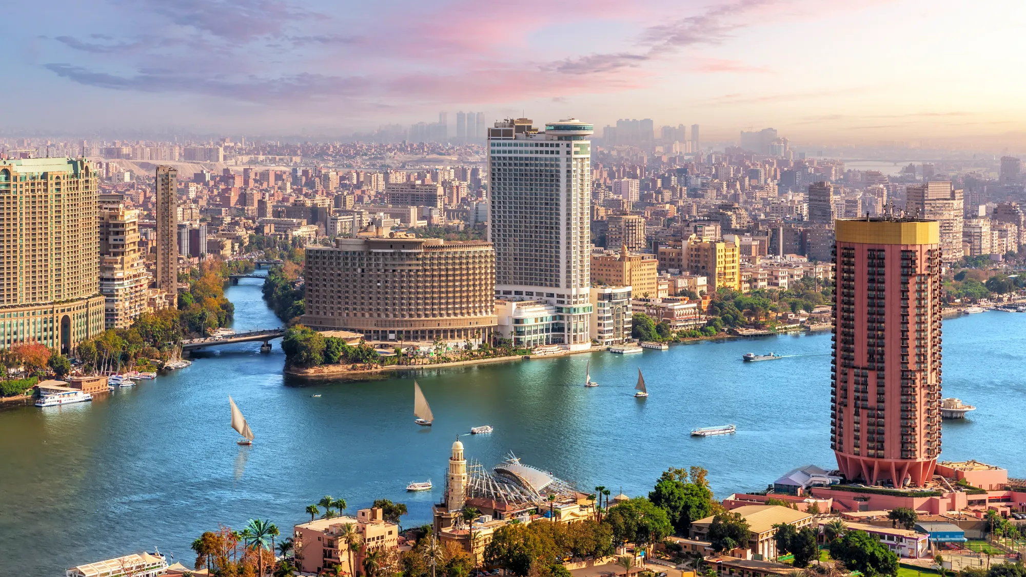 Aerial view of downtown Cairo with the Nile River, high-rise buildings, boats on the water, and a hazy skyline under a pink and blue sky at sunset.